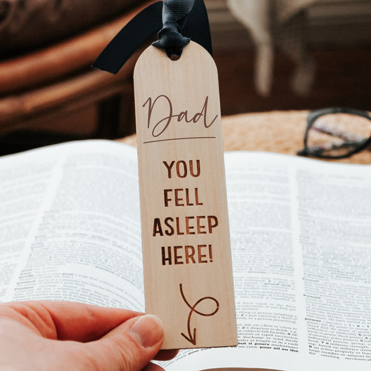 Playful wooden bookmark engraved with "Dad you fell asleep here" message pointing to a book below. Finished with a blue ribbon, it's the perfect reminder for book-loving fathers.