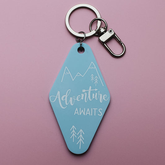 Blue motel style keyring tag with 'Adventure Awaits' text on a pink background