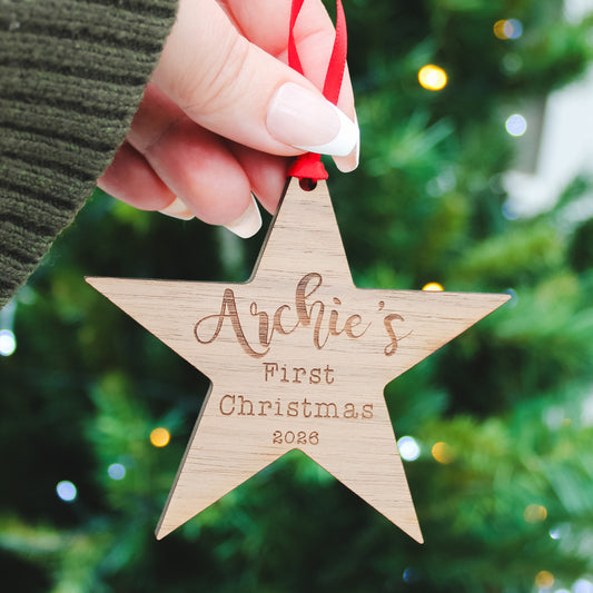 A hand holds a personalized wooden star Christmas ornament engraved with 'Archie's First Christmas 2026' in a cosy holiday setting with a blurred Christmas tree in the background.