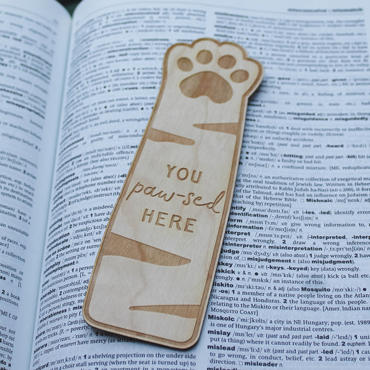 Wooden bookmark with paw design and 'You paw-sed here' text engraved onto it, sitting on an open book.