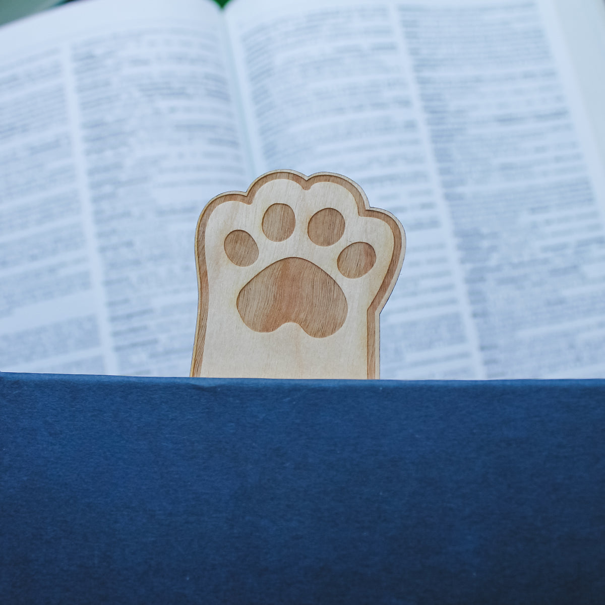 Wooden paw print bookmark sitting inside blue book so the paw is sticking out the top with an open book in the background