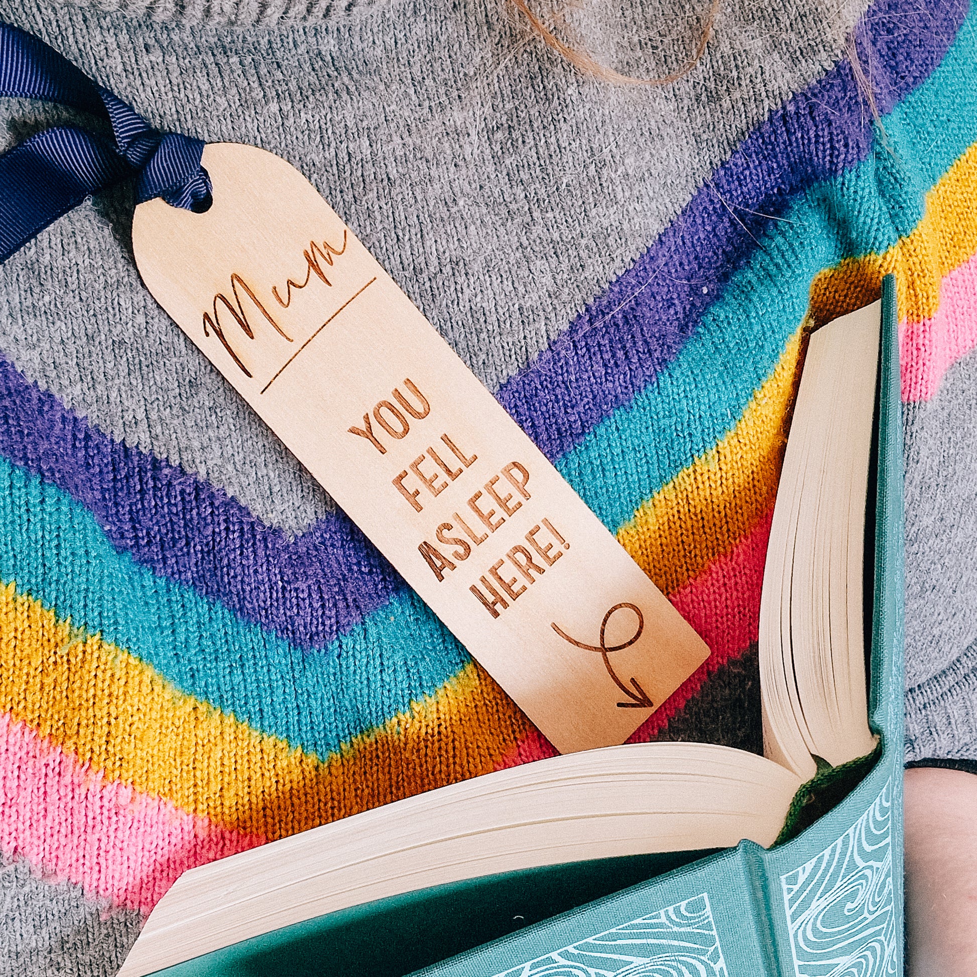 Personalized wooden bookmark engraved with "Mum, you fell asleep here" and an arrow pointing to the bottom. The bookmark rests on a person sleeping with an open book with a navy blue ribbon attached.