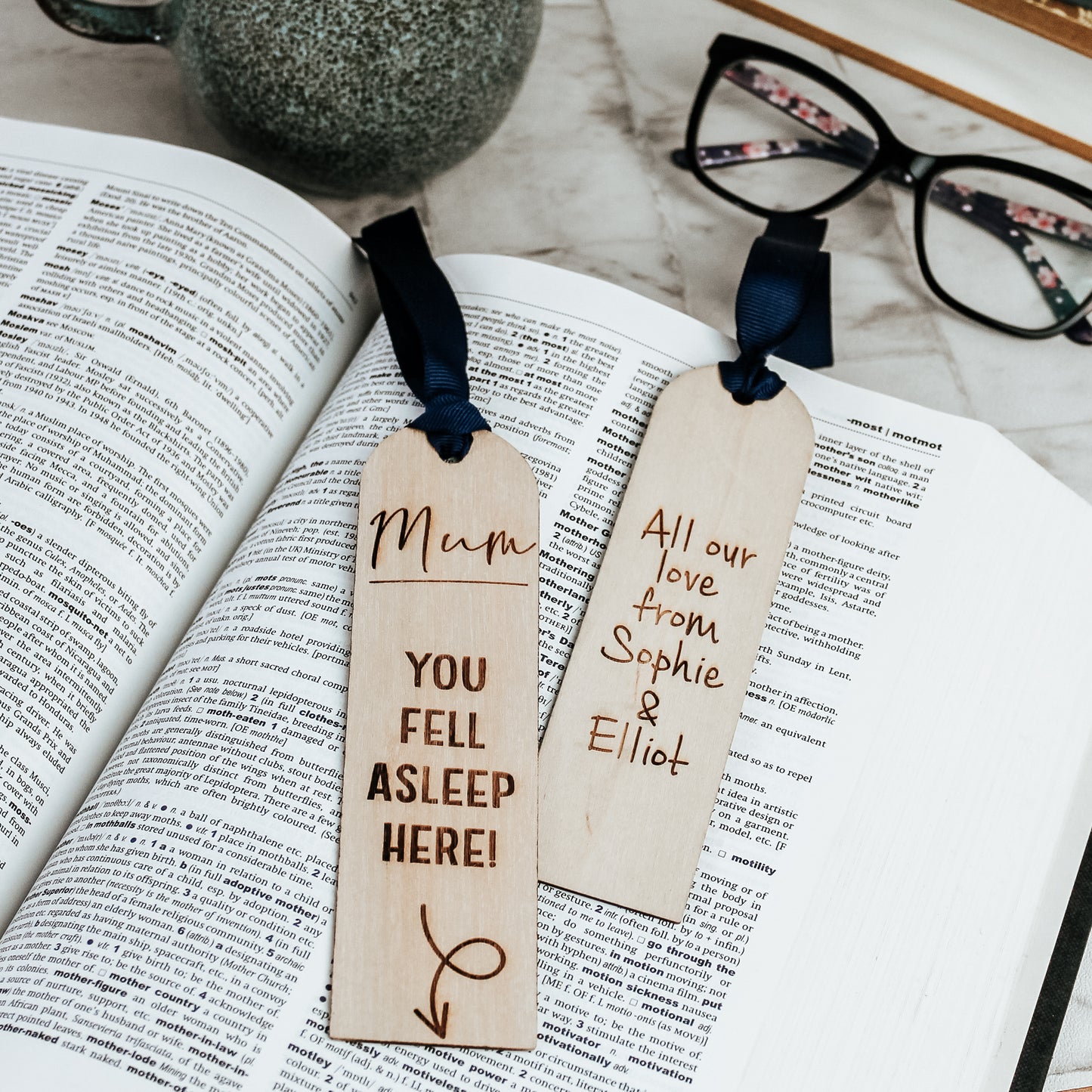 Personalised wooden bookmark engraved with "Mum, you fell asleep here" and an arrow pointing to the bottom. The bookmark rests on an open book with a navy blue ribbon attached.