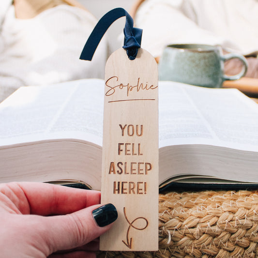 Engraved wooden bookmark personalized with the you own name and the playful message "you fell asleep here" with an arrow pointing to the book below. Finished with a blue ribbon, this bookmark adds a touch of whimsy to any reading experience