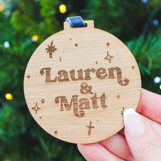 Wooden Christmas bauble ornament with personalised names engraved, held against a blurred green background.