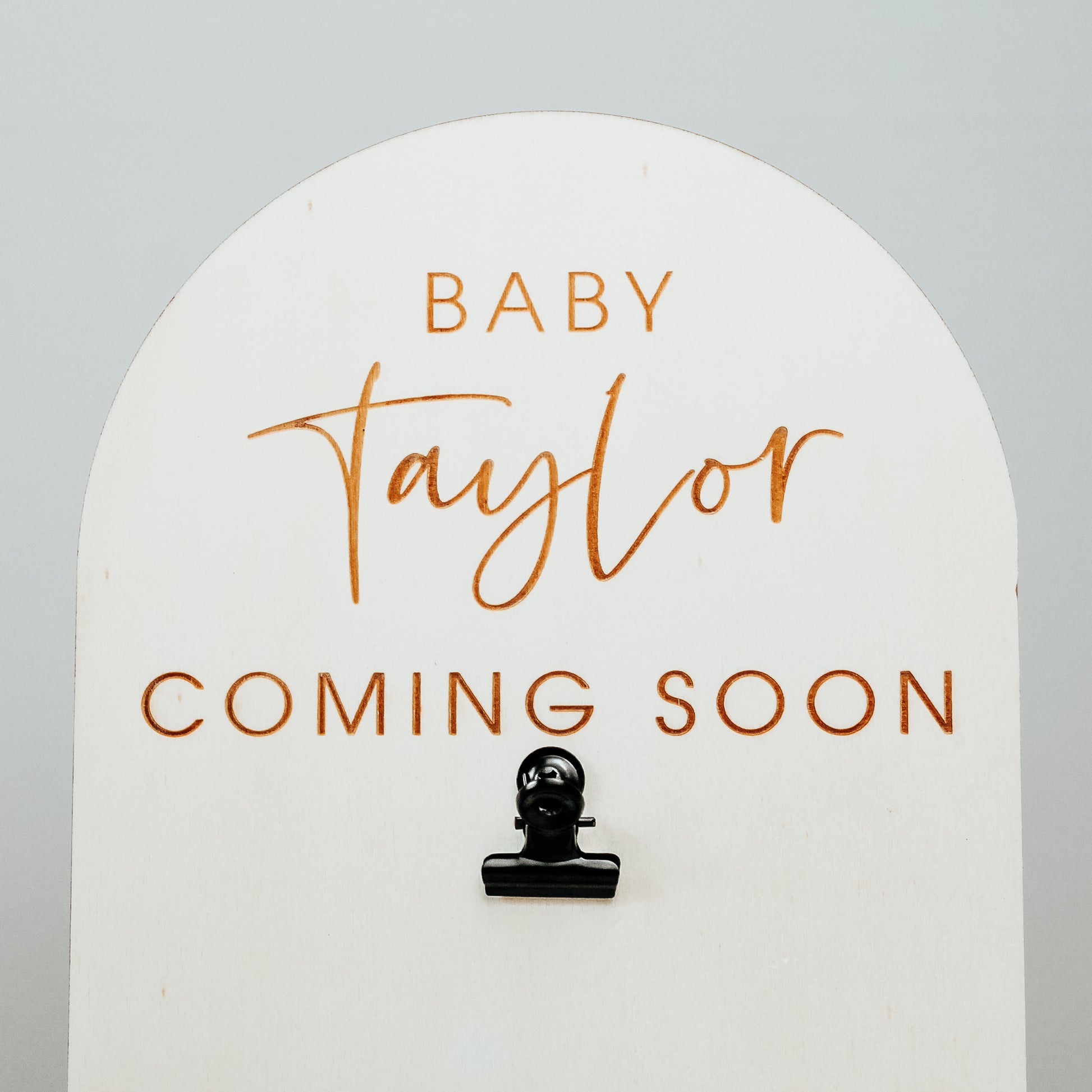 image of the top of the pregnancy announcement sign showing the engraved personalised wording close up. It reads baby taylor coming soon and has a black clip to clip the baby scan image onto