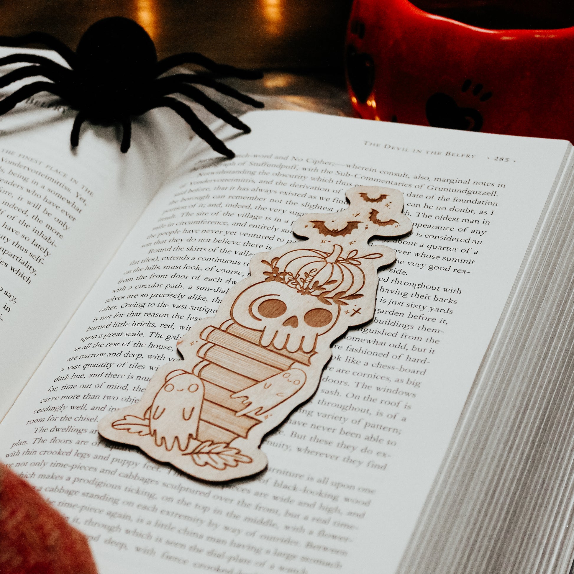 Wooden engraved bookmark with halloween design, including a pumpkin, skull and ghosts, placed on a book with spooky decorations placed around