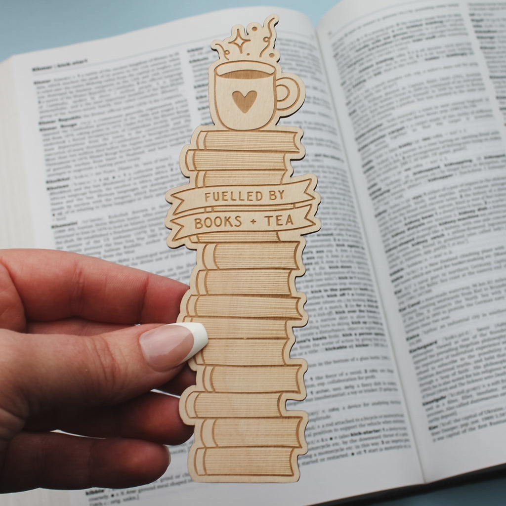 Wooden bookmark with 'Fuelled by Books + Tea' design held over an open book.