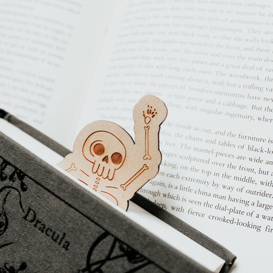 Skeleton bookmark, made from wood and engraved with a skeleton design, peeking their head out of a Dracula horror book and waving