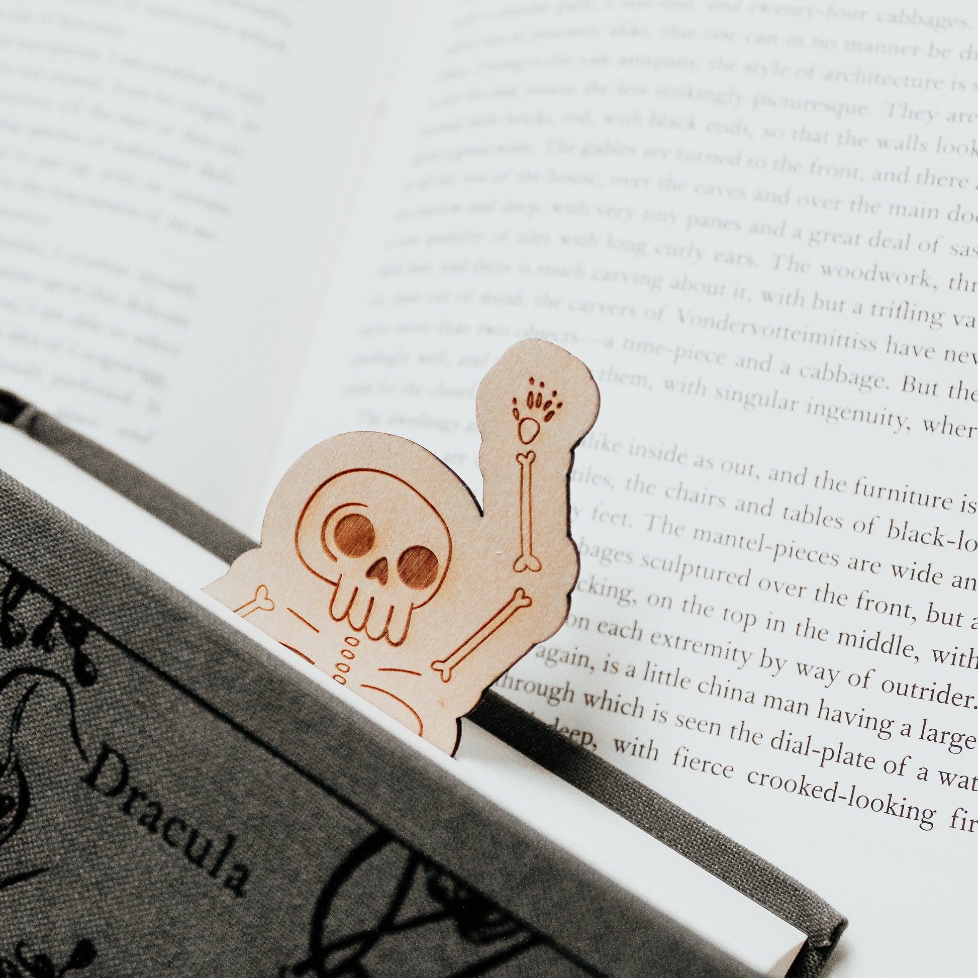 Skeleton bookmark, made from wood and engraved with a skeleton design, peeking their head out of a Dracula horror book and waving 