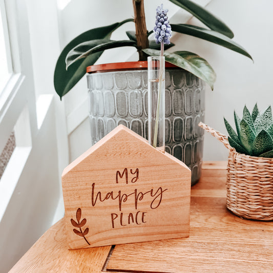 wooden house shaped block with test tube coming out of the top, holding a cut flower, engraved with the wording my happy place