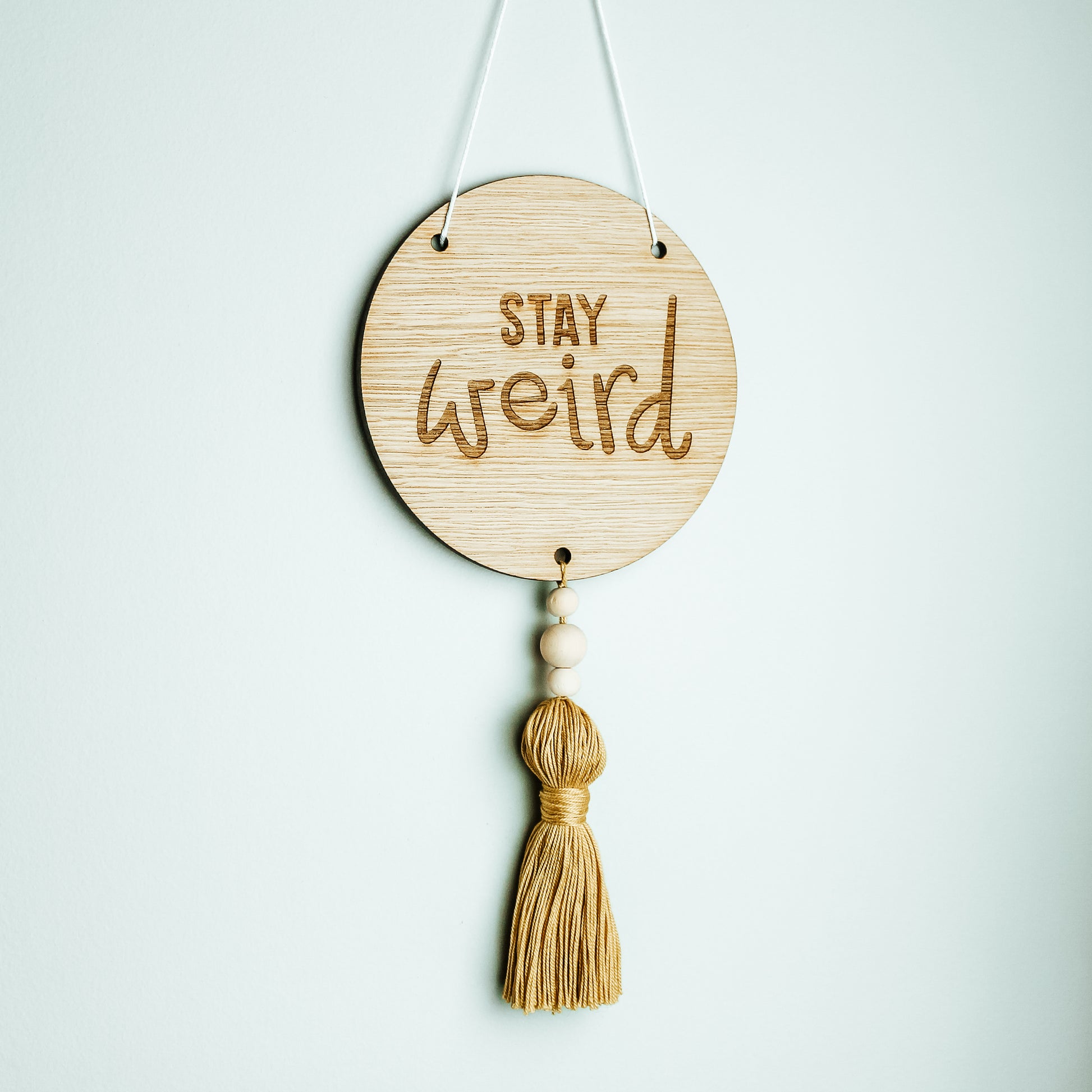 round wooden motivational sign with the words stay weird on it affixed to a yellow tassel 