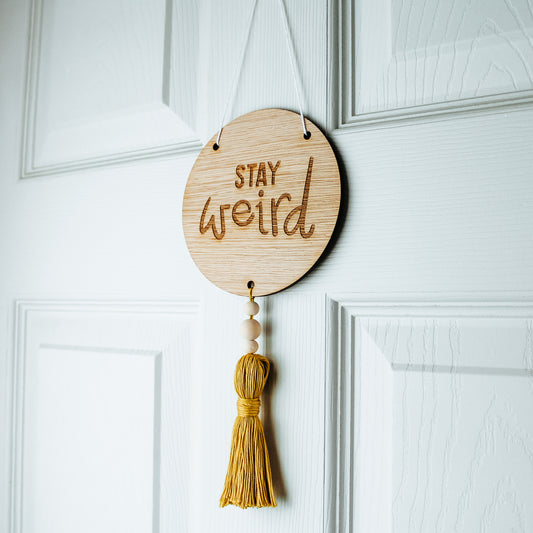 round wooden sign engraved with the phrase stay weird with a coloured tassel affixed to the bottom