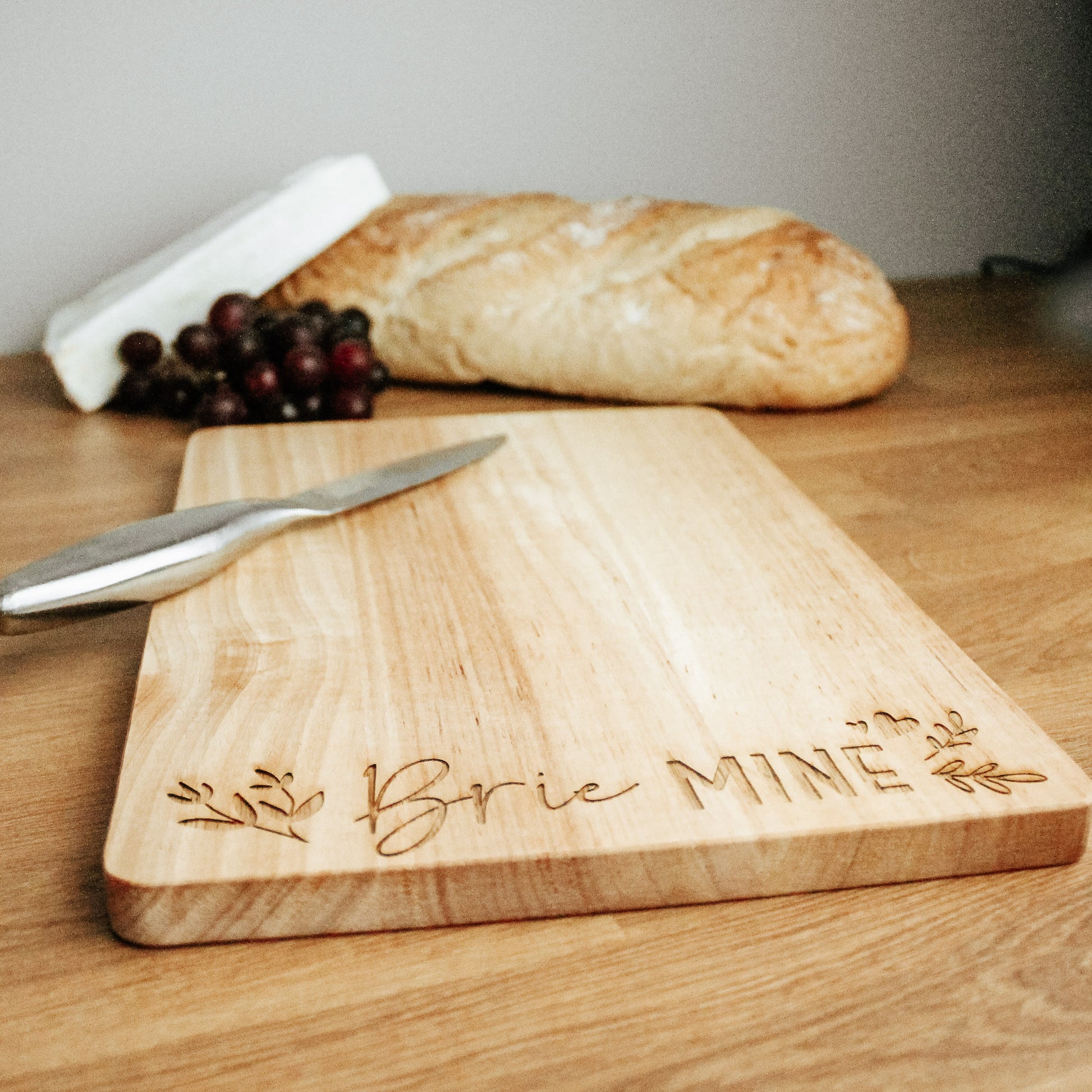 Cheese serving board in a light wood, with the words brie mine in a calligraphy font engraved on it, with some floral design on the side. A wedge of brie, bread and grapes can be seen in the background and a knife on top of the board