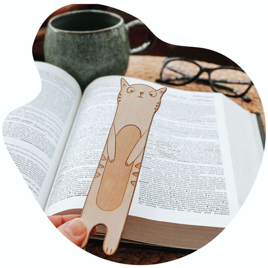 image of a wooden engraved cat shaped bookmark held before an open book and a mug 
