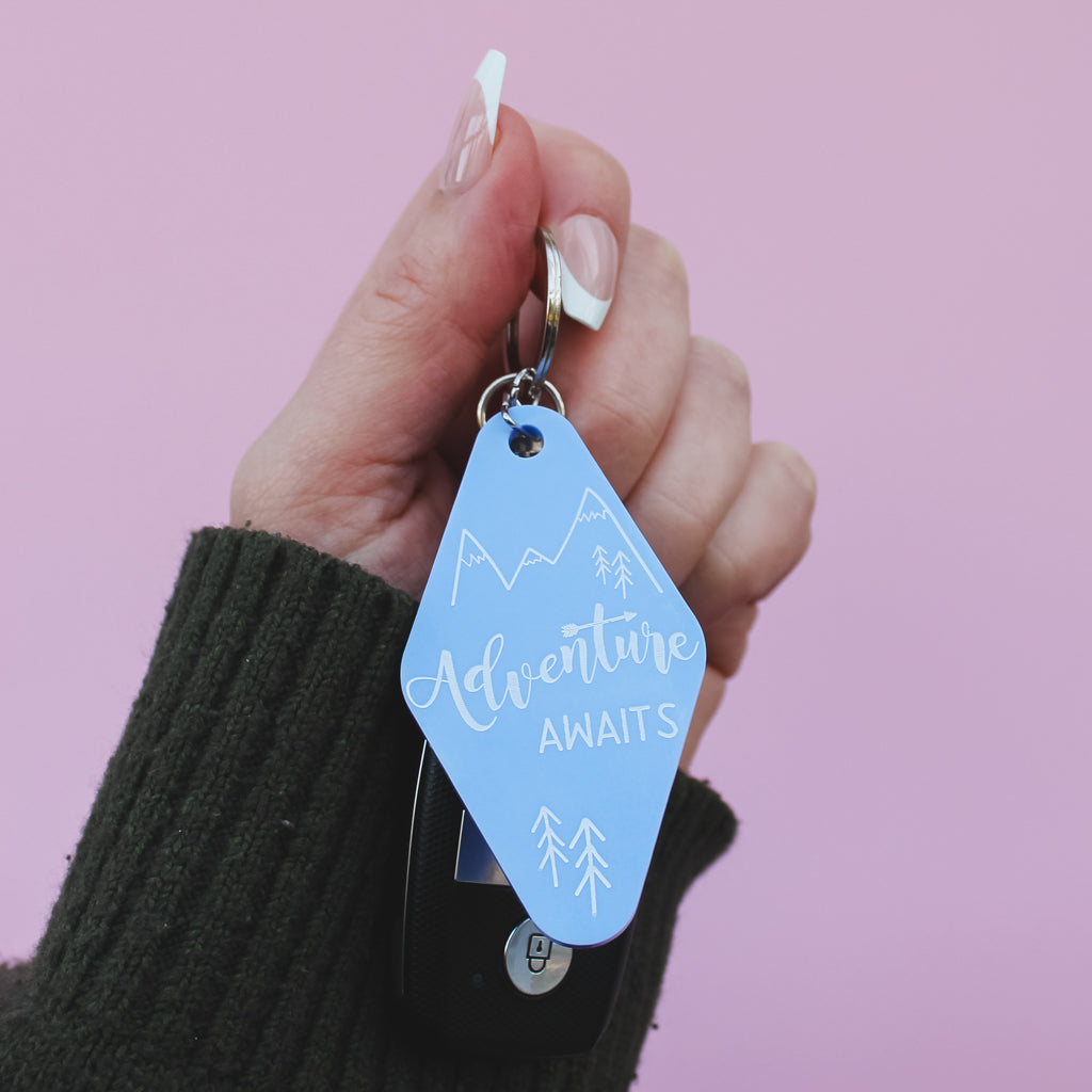Hand holding a blue motel style keyring with 'Adventure Awaits' text against a pink background