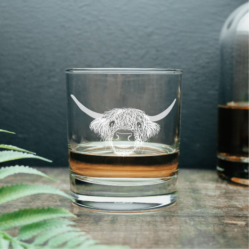 Hand drawn Highland cow design, engraved on a clear whisky glass tumbler with a wee dram of whisky inside of it. 