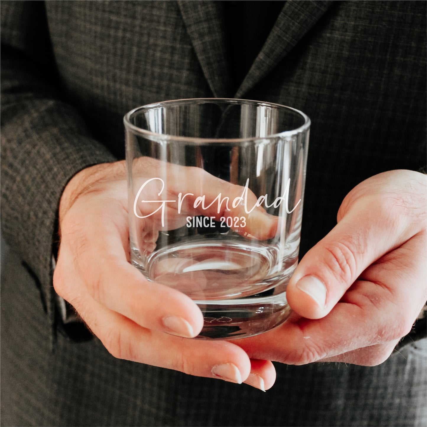 man holding a whisky tumbler glass engraved with the phrase Grandad since 2023 