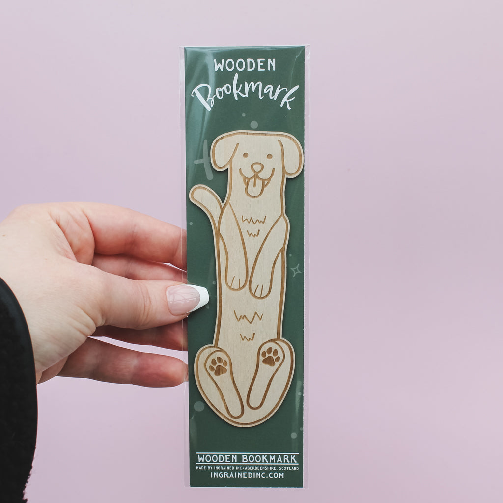 Hand holding a wooden bookmark in its packaging with a dog design against a light pink background