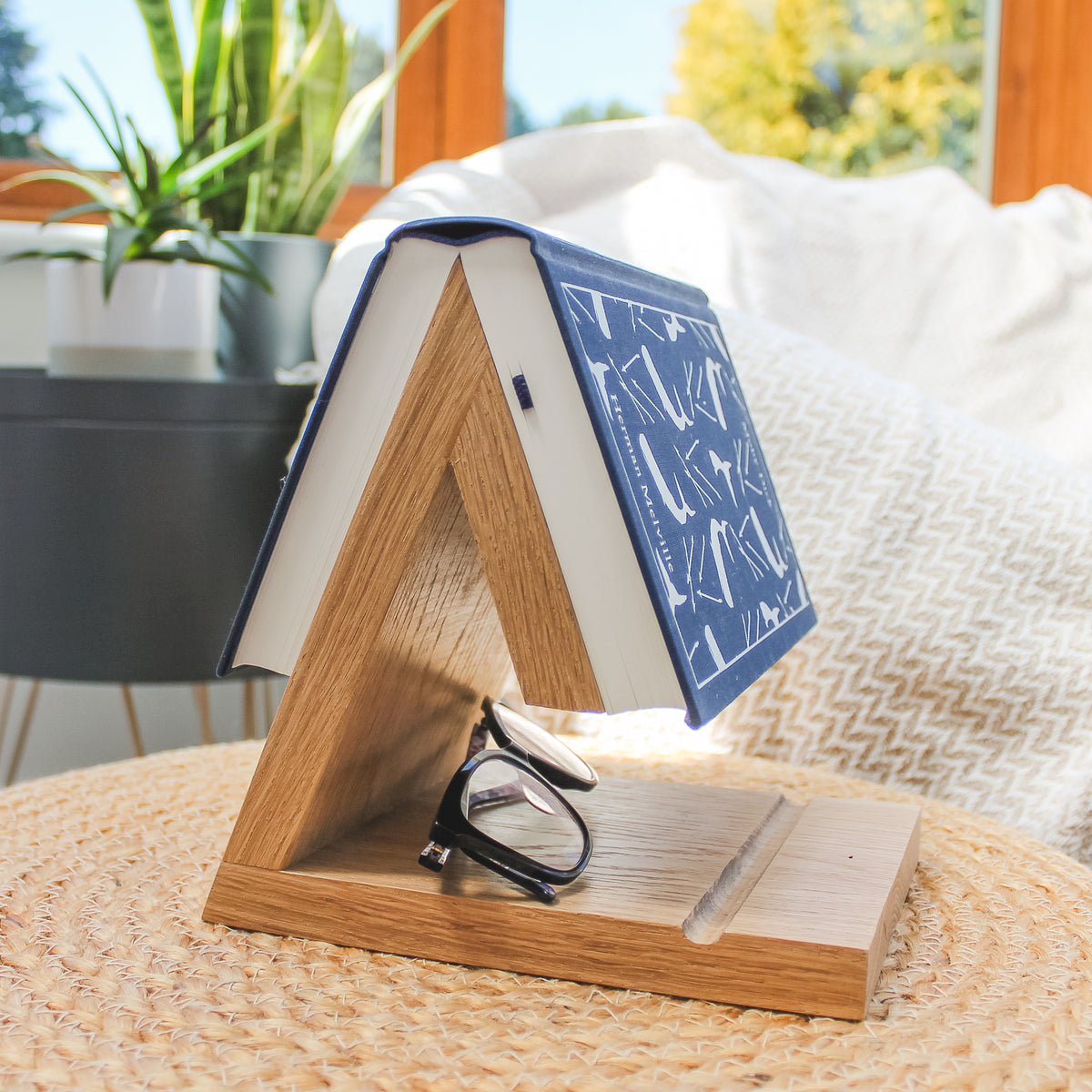 A warm wooden oak book stand holding an open book and a pair of eyeglasses, placed on a woven table in a bright living room setting.