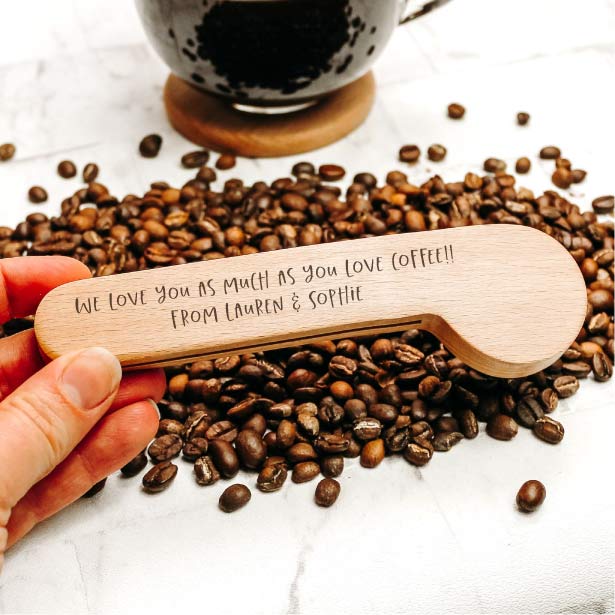 a wooden coffee scoop with personalised engraving on the reverse with a bed of coffee in the background 