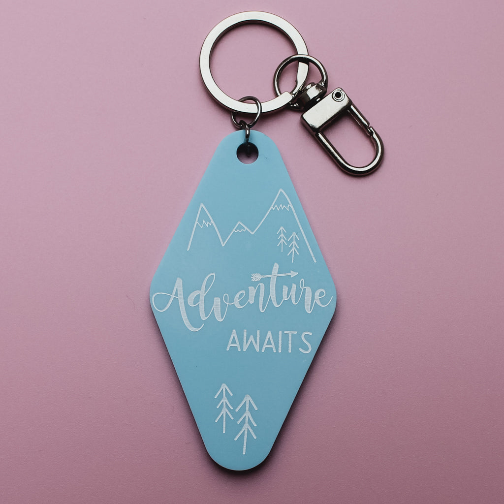 Blue motel style keyring tag with 'Adventure Awaits' text on a pink background