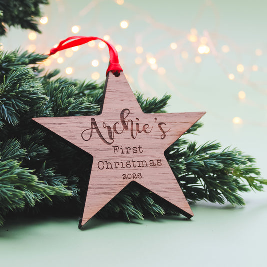 A wooden five-point star Christmas ornament engraved with a personalised name First Christmas 2026' and hanging from a red ribbon, displayed among green pine branches against a soft, blurred holiday background.