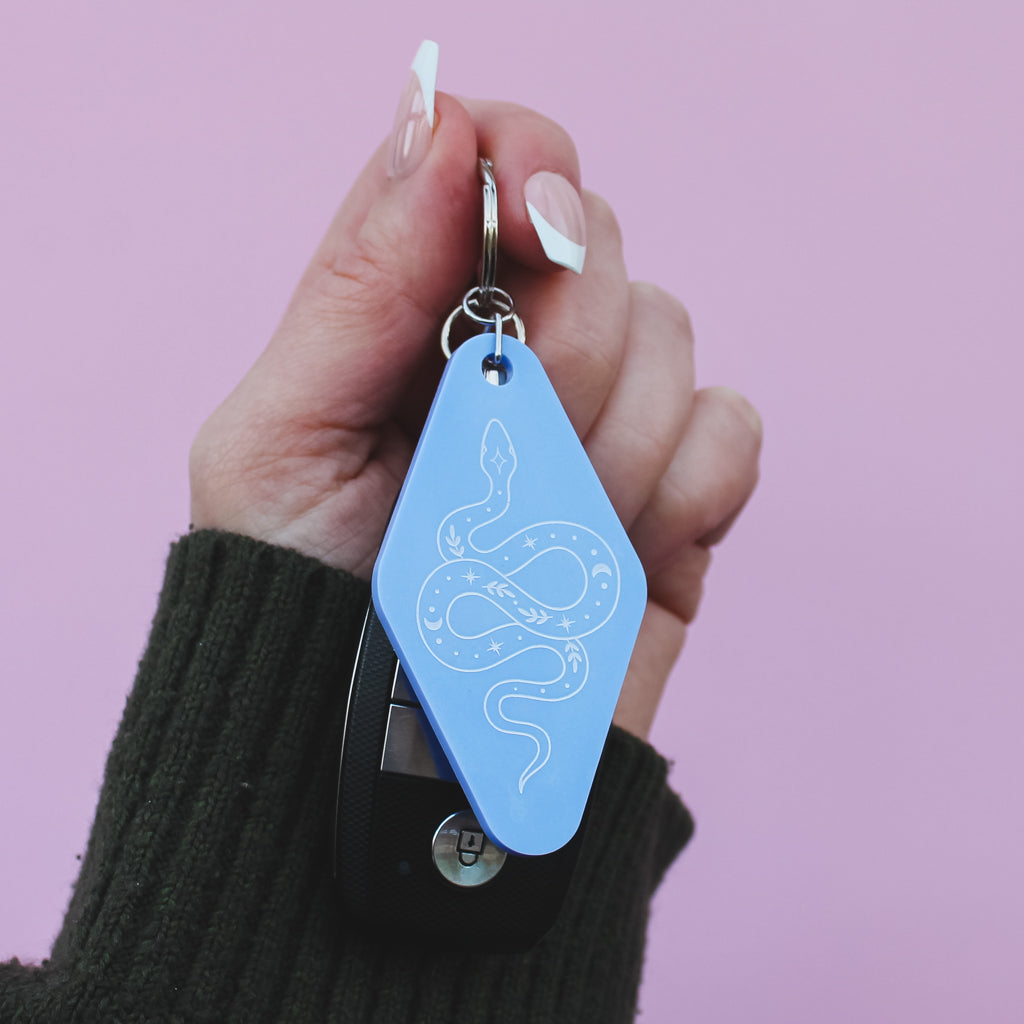 Hand holding a blue motel style 
keychain with a snake design against a pink background