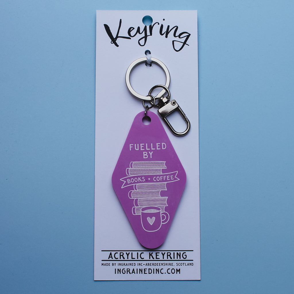 Pink acrylic keyring with text saying, 'fuelled by books and coffee' on a white  backing card against a blue background