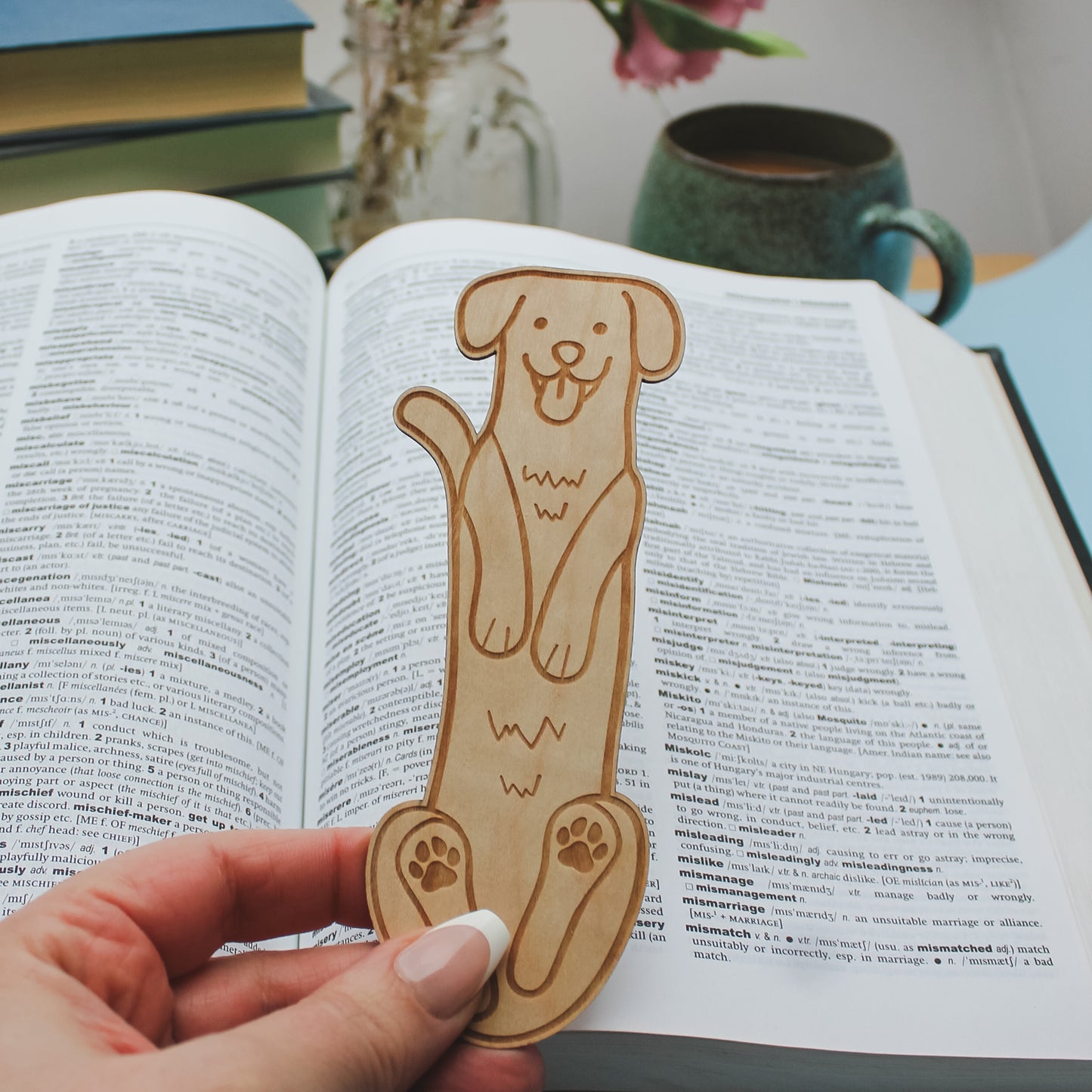 Dog-shaped wooden bookmark held over an open book with a cup and books in the background.
