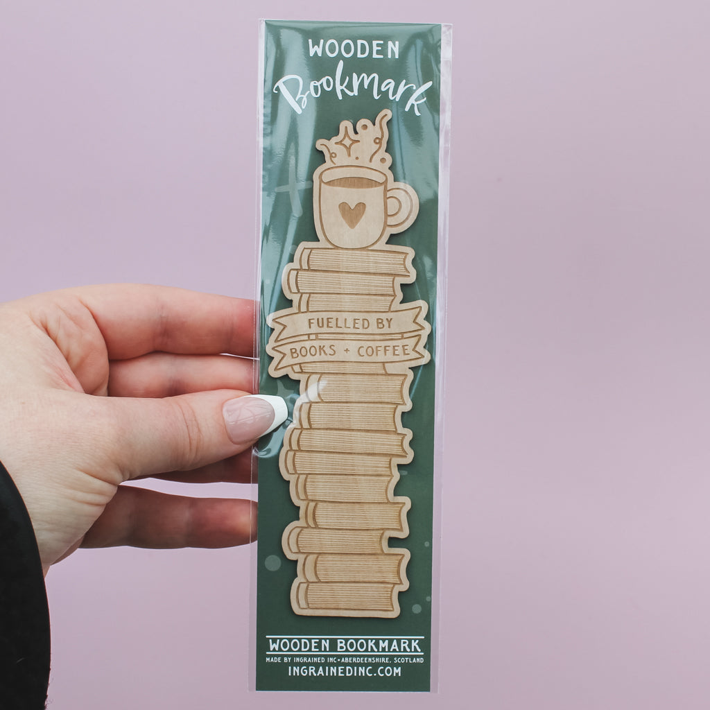 Hand holding a wooden packaged bookmark with a design of books and a coffee cup on a pink background
