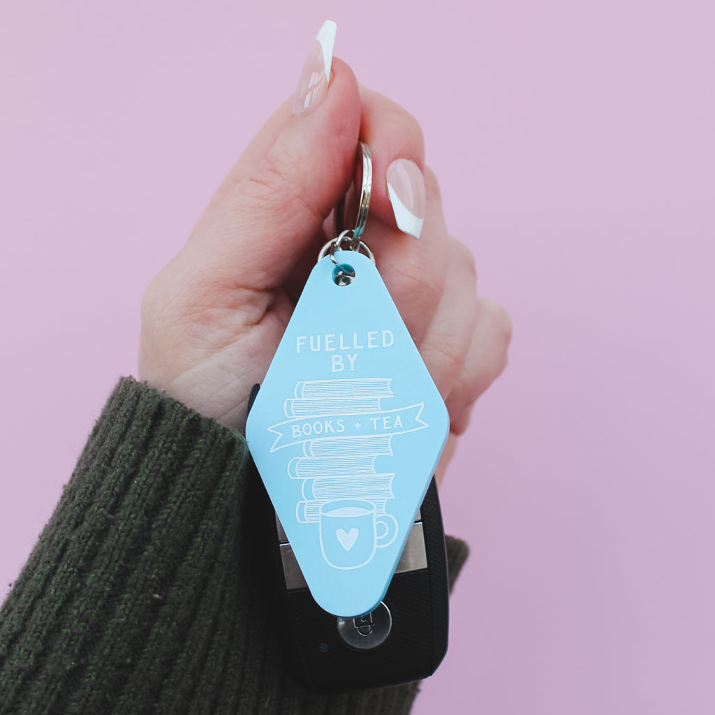 Hand holding a blue motel style keychain with 'Fuelled by Books & Tea' text against a pink background