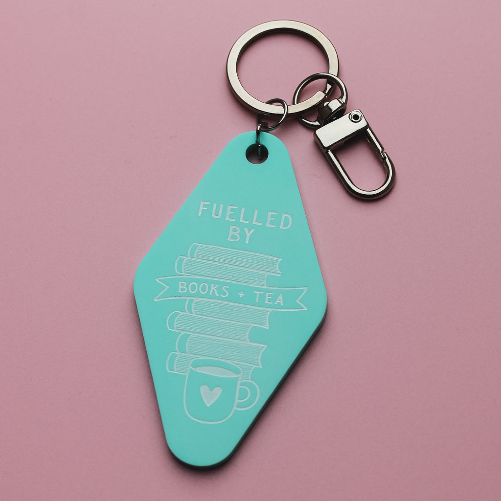 Turquoise motel style keychain with 'Fuelled by Books + Tea' text on a pink background