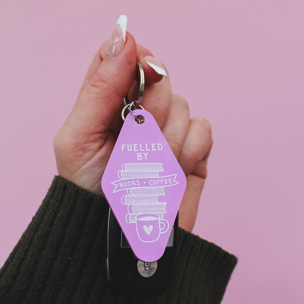 Hand holding a pink motel style keychain with 'Fuelled by Books & coffee' text against a pink background