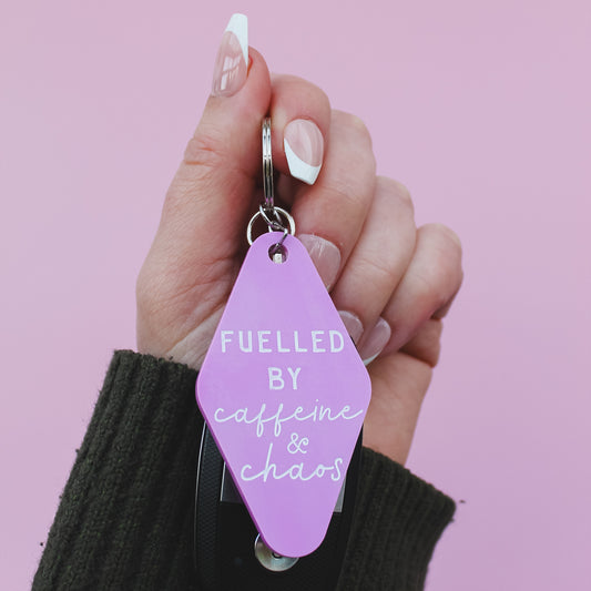 Hand holding a pink motel style keychain with 'Fuelled by caffeine & chaos' text on a pink background