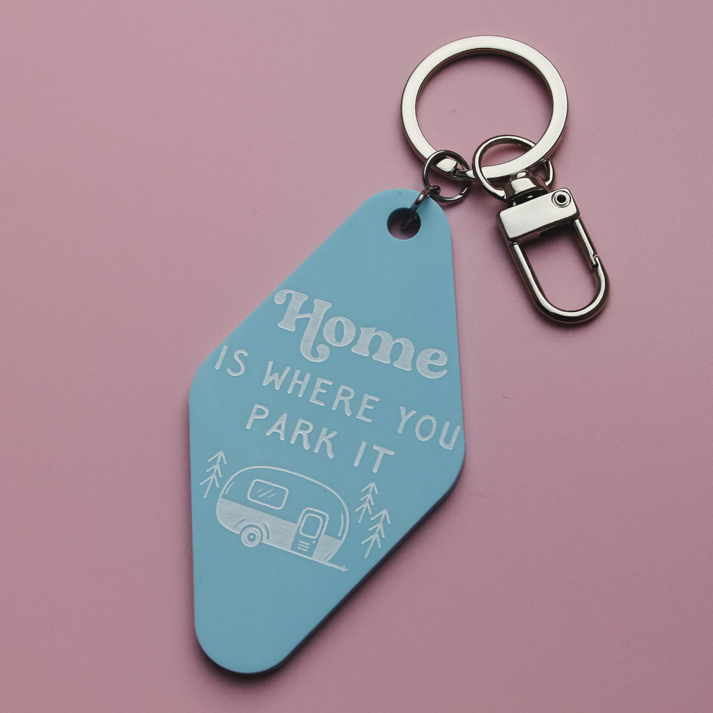 Blue motel style keychain with 'Home is where you park it' text and caravan illustration on a pink background