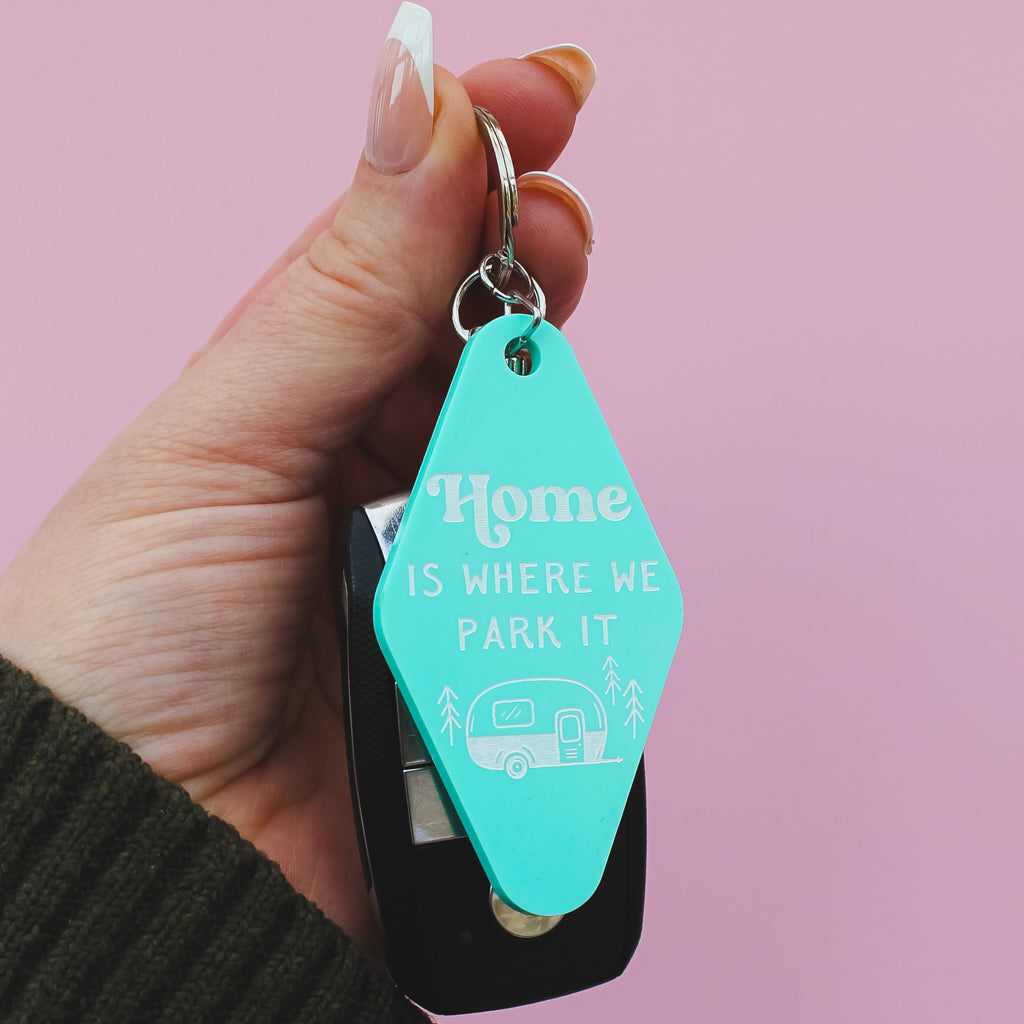 Turquoise motel style keychain with 'Home is where we park it' text and a black clip on a pink background.