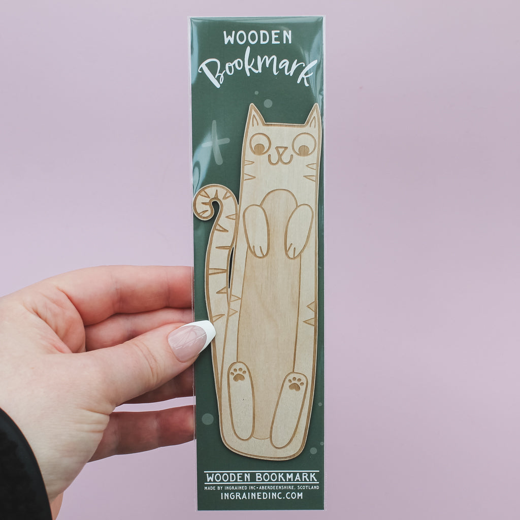Hand holding a wooden bookmark in packaging shaped like a cat against a light pink background