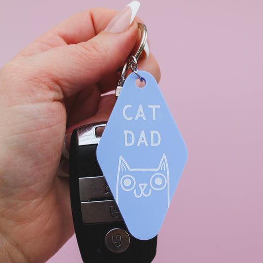 Hand holding a blue Motel Style keychain with 'CAT DAD' tag against a pink background
