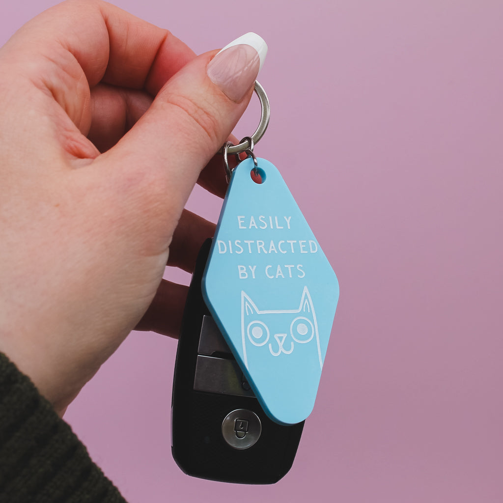 Hand holding a blue motel style keychain with 'easily distracted by cats' engraved on it and cat illustration against a pink background