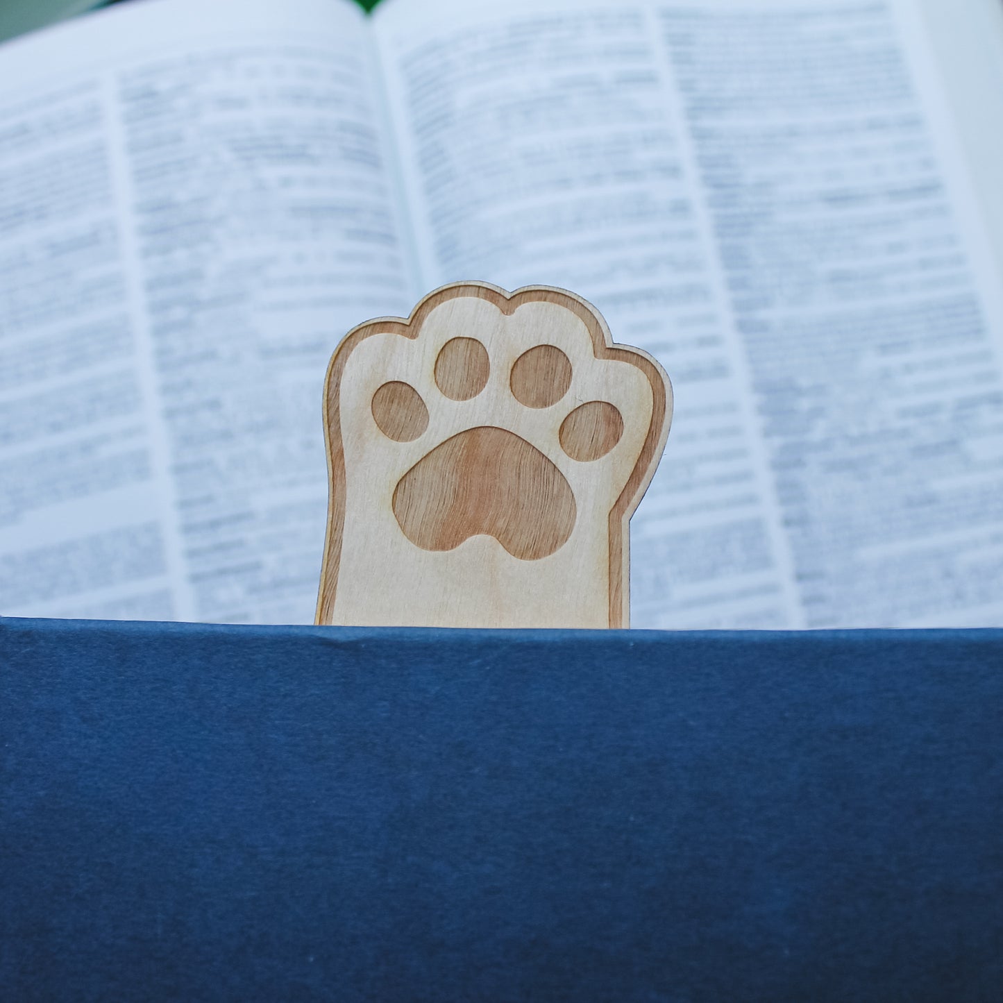 Wooden paw print bookmark sitting inside blue book so the paw is sticking out the top with an open book in the background 