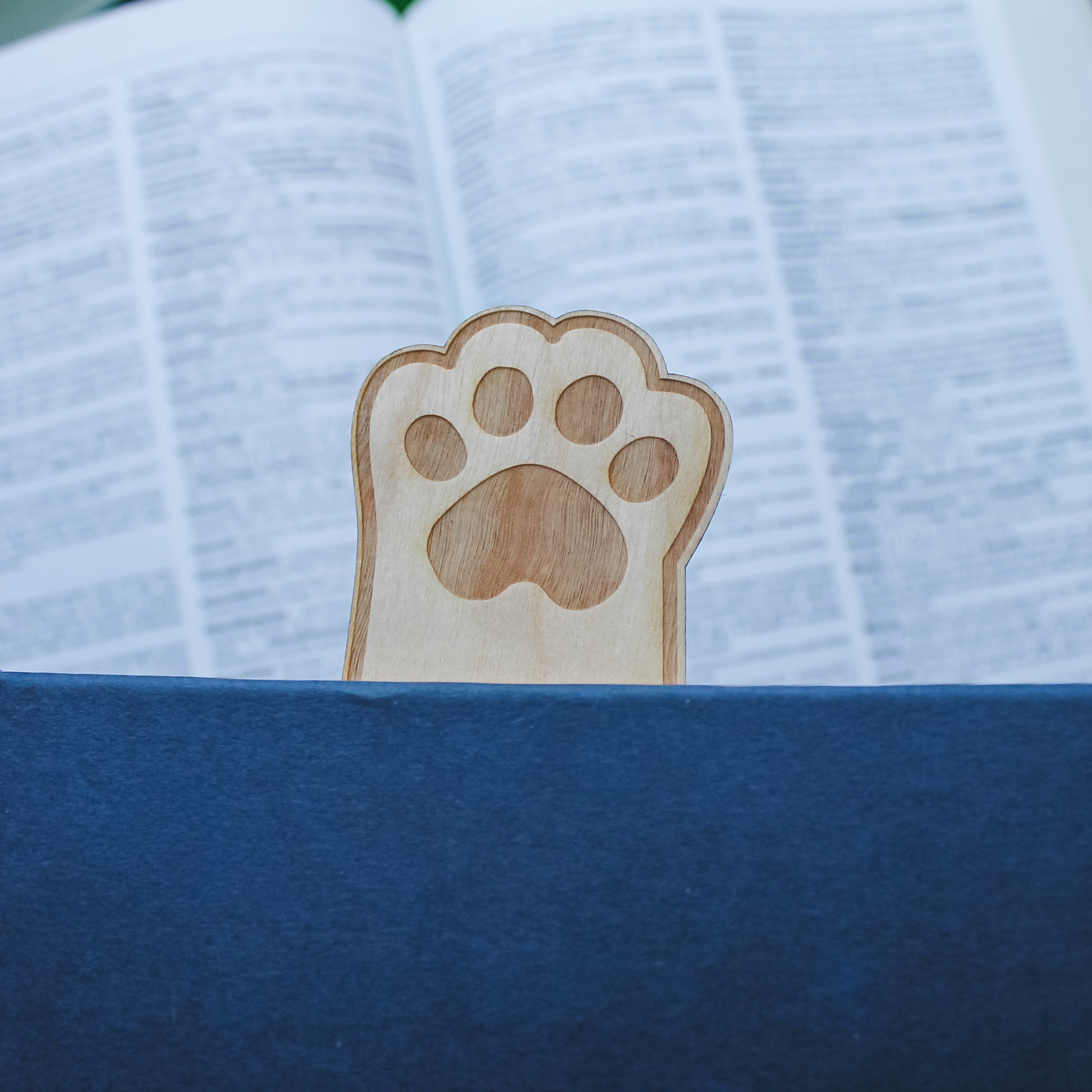 Wooden paw print bookmark sitting inside blue book so the paw is sticking out the top with an open book in the background 