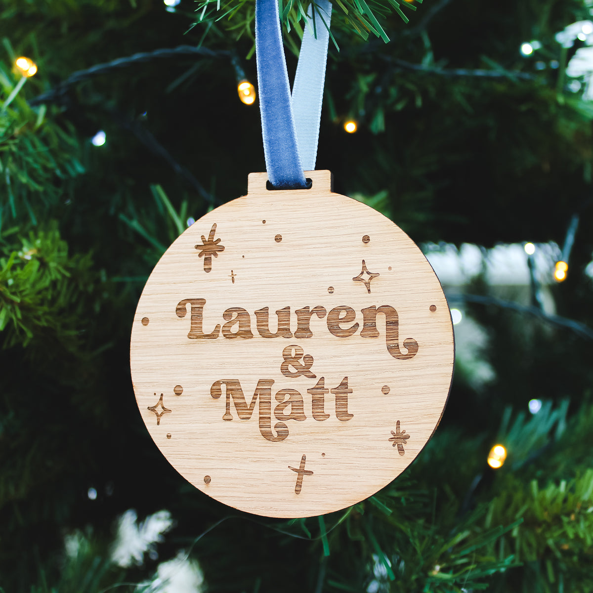 Wooden modern christmas bauble ornament with a custom couples name  engraved, hanging on a Christmas tree.