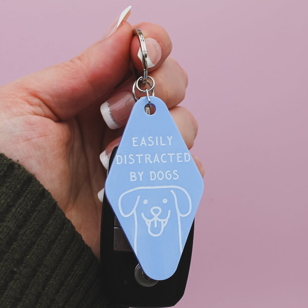 Hand holding a blue motel style  keychain with 'Easily Distracted by Dogs' text against a pink background