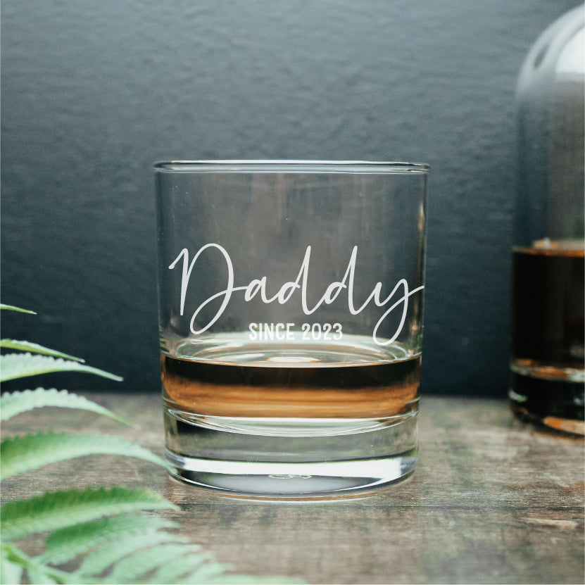 Image of a whisky glass with the wording daddy since 2023 engraved onto it. Filled slightly with a dram of whisky.
