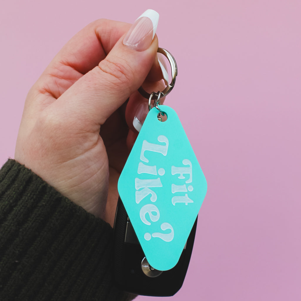 Hand holding a turquoise motel style keyring with 'Fit Like?' text against a pink background