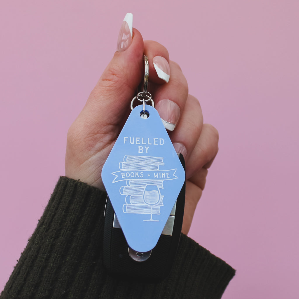 Hand holding a blue motel style keychain with 'Fuelled by Books + Wine' text against a pink background