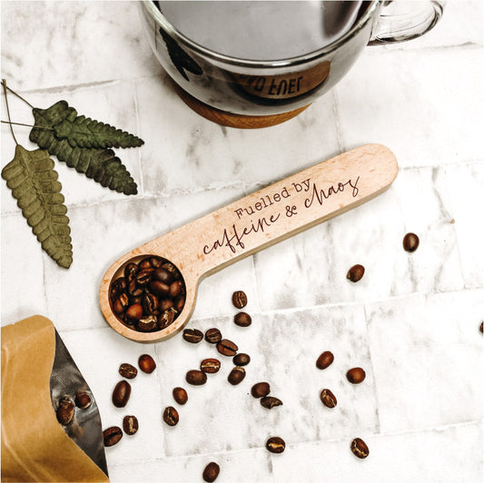 wooden coffee spoon engraved with the words fuelled by caffeine and chaos surrounded by coffee accoutrements and coffee beans