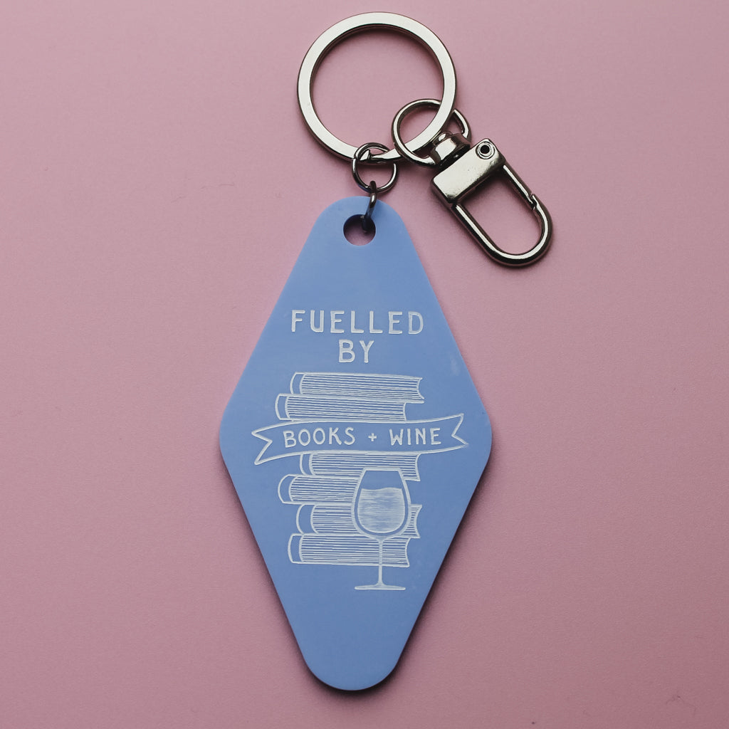 Blue motel style keychain with 'Fuelled by Books + Wine' text on a pink background