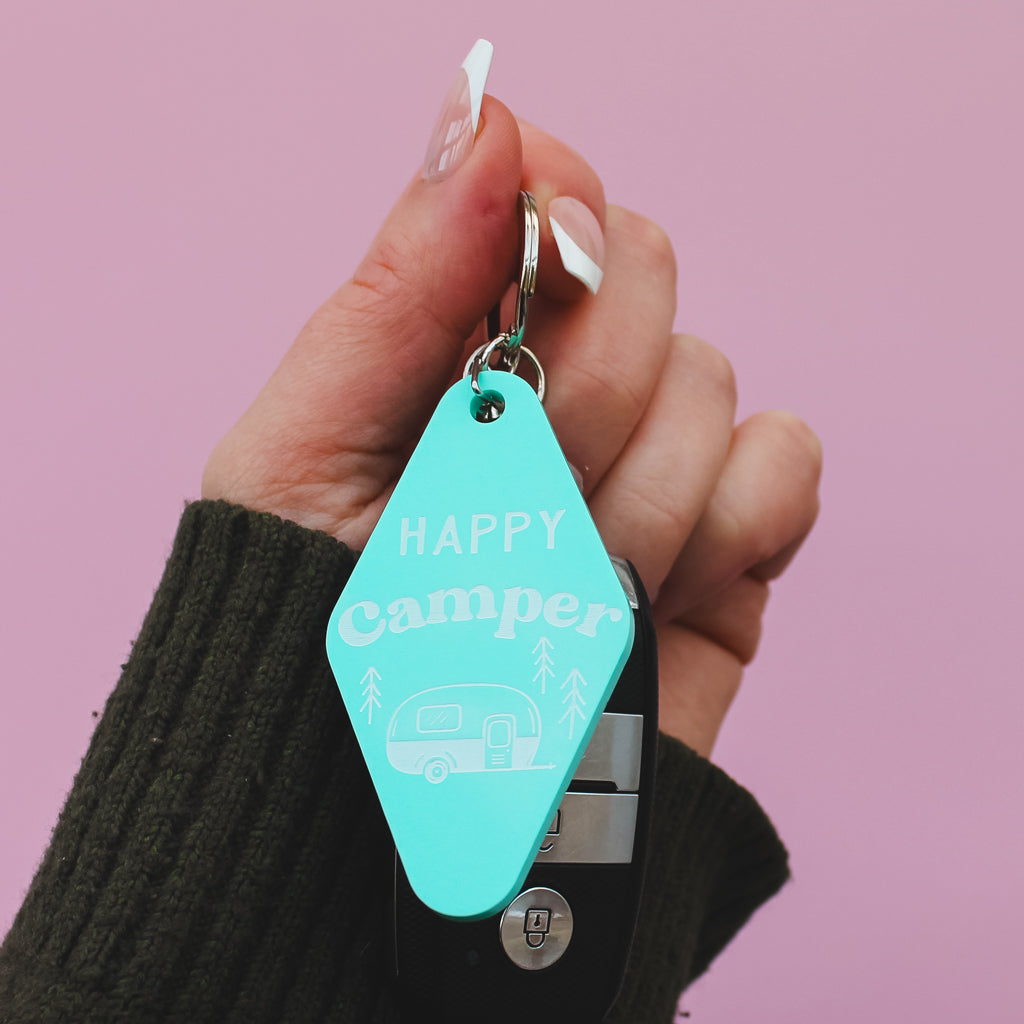 Turquoise motel style keychain with 'Happy Camper' text held by a hand against a pink background